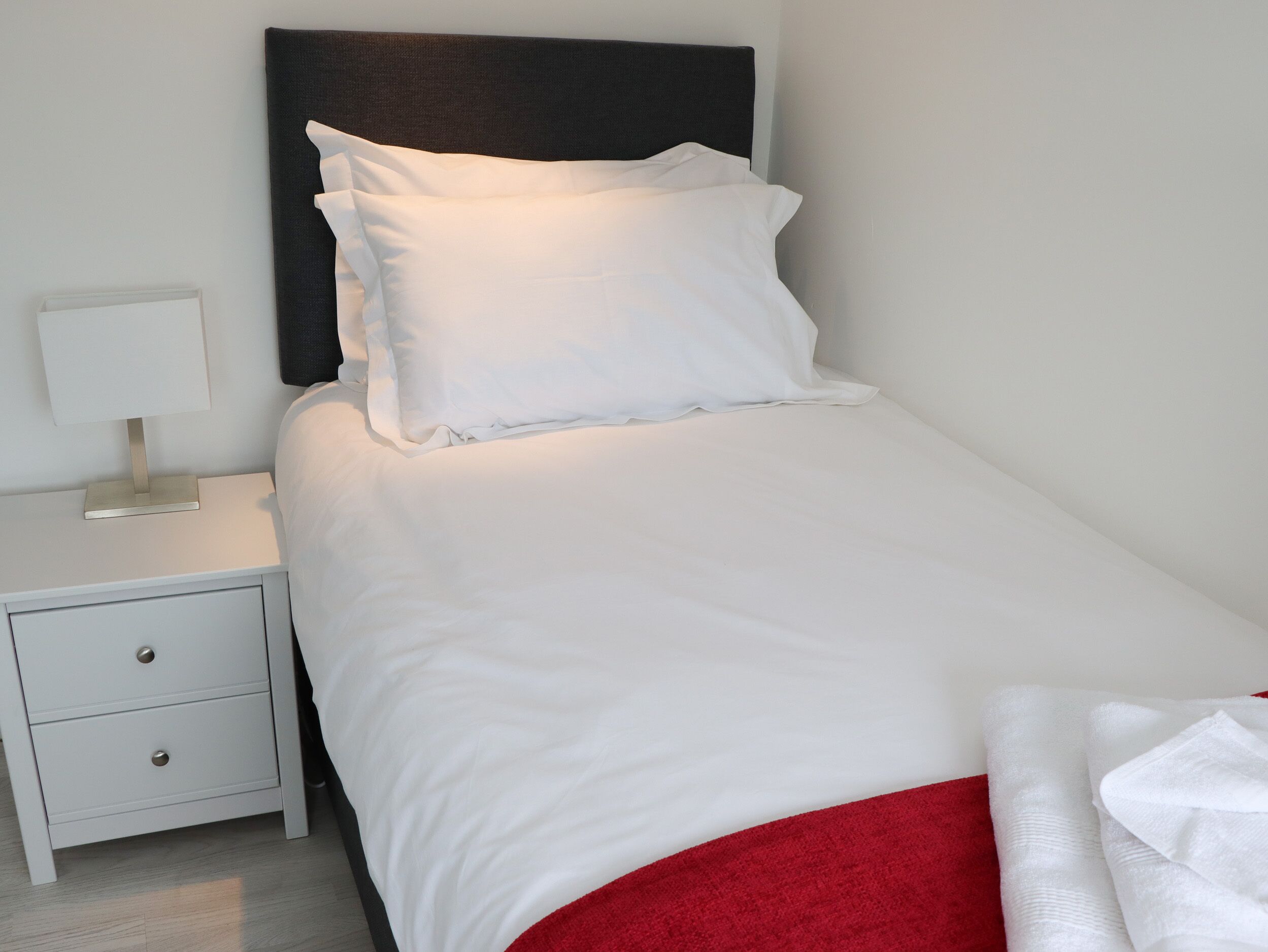 Un lit bien fait, avec du linge de lit blanc et une couverture rouge, une table de chevet blanche avec une lampe, et une tête de lit sombre.