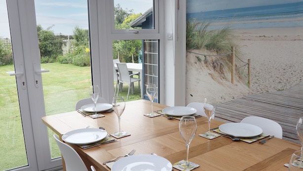 A dining table set for six with white plates and glasses, positioned near a large window overlooking a beach.