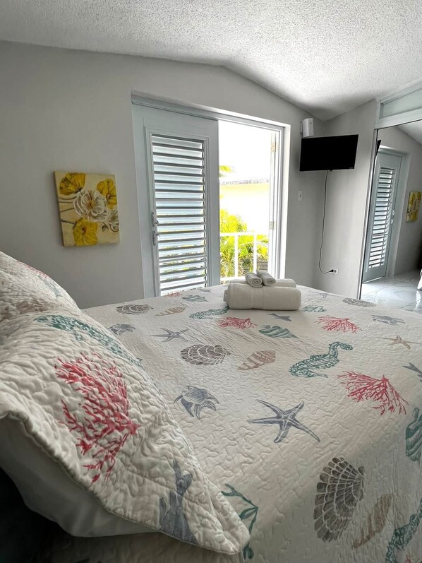 A bedroom with a bed, a painting on the wall, a TV mounted on the wall, and a window with blinds.