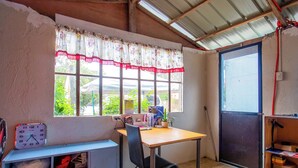 A small, rustic room with a wooden desk, a chair, a window with a floral curtain, a clock, and a shelf with various items. - OYO 814 Laurien's Budget Hotel (Tagaytay)