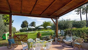 Une terrasse extérieure dotée d’une pergola en bois, de chaises en plastique blanches, d’une table dressée pour le repas, et offrant une vue sur un espace verdoyant agrémenté de parasols et de chaises au fond.