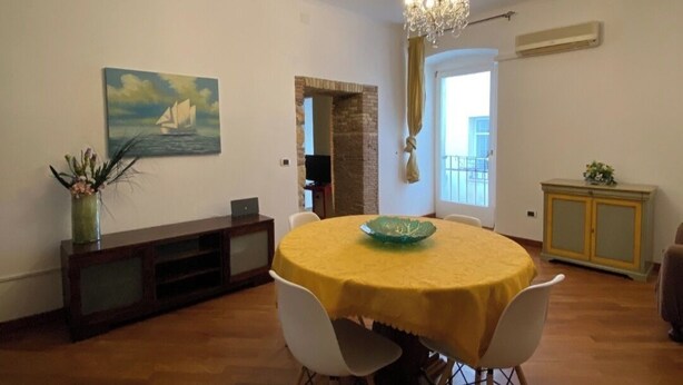 Un comedor con una mesa redonda cubierta de amarillo, rodeada de sillas blancas. Hay un cuadro en la pared, una lámpara de araña y un aparador con una planta.