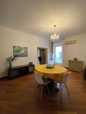 Ein Essbereich mit einem runden Tisch, der gelb bedeckt ist, umgeben von weißen Stühlen. An der Wand hängt ein Gemälde, eine Chandelaber und ein Schrank mit einer Pflanze.