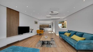 Una sala de estar moderna con televisor de pantalla plana, un sofá azul con cojines amarillos, piso de madera y una cocina al fondo.