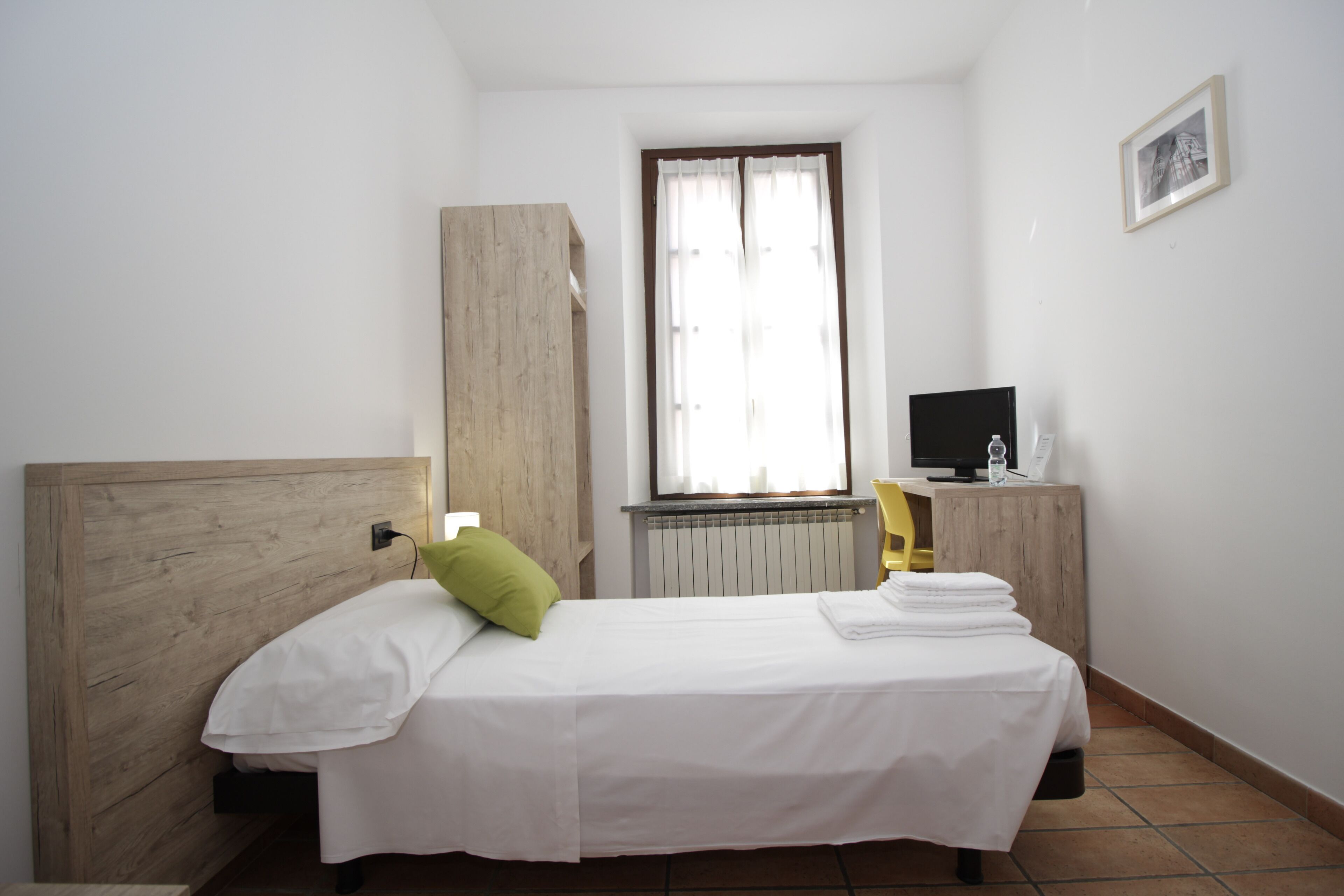 A bedroom with a wooden headboard, a bed with white bedding, a green pillow, a radiator, a television on a wooden stand, and a framed picture on the wall.