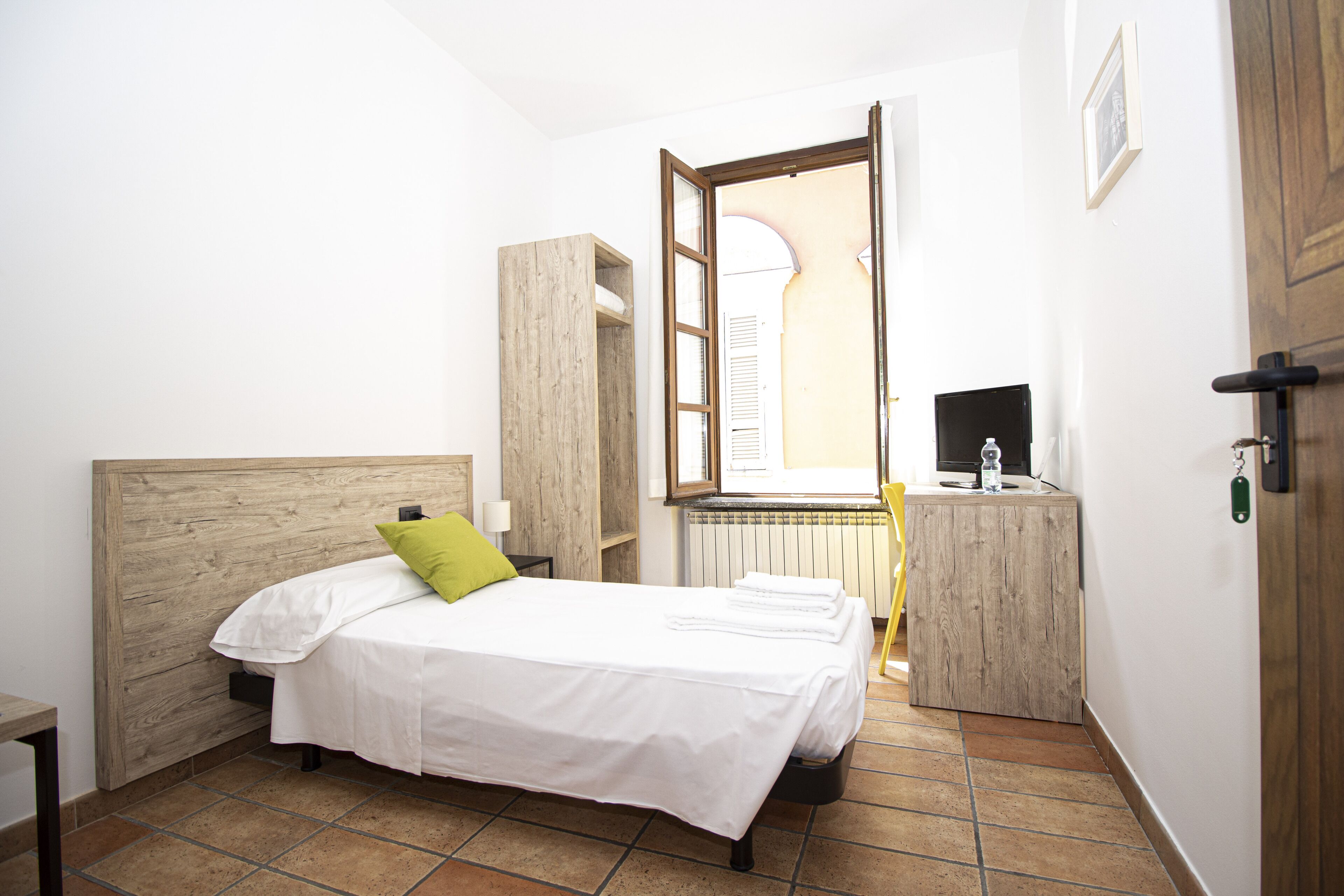 Een slaapkamer met een houten hoofdbord, een bed met witte lakens en een groen kussen, een nachtkastje, een klein bureau met een computer, en een raam met uitzicht op een gebouw.