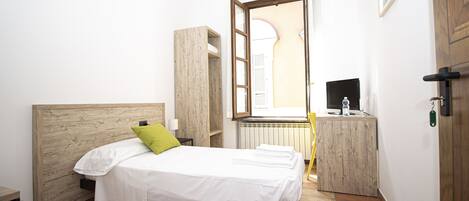 Een slaapkamer met een houten hoofdbord, een bed met witte lakens en een groen kussen, een nachtkastje, een klein bureau met een computer, en een raam met uitzicht op een gebouw.