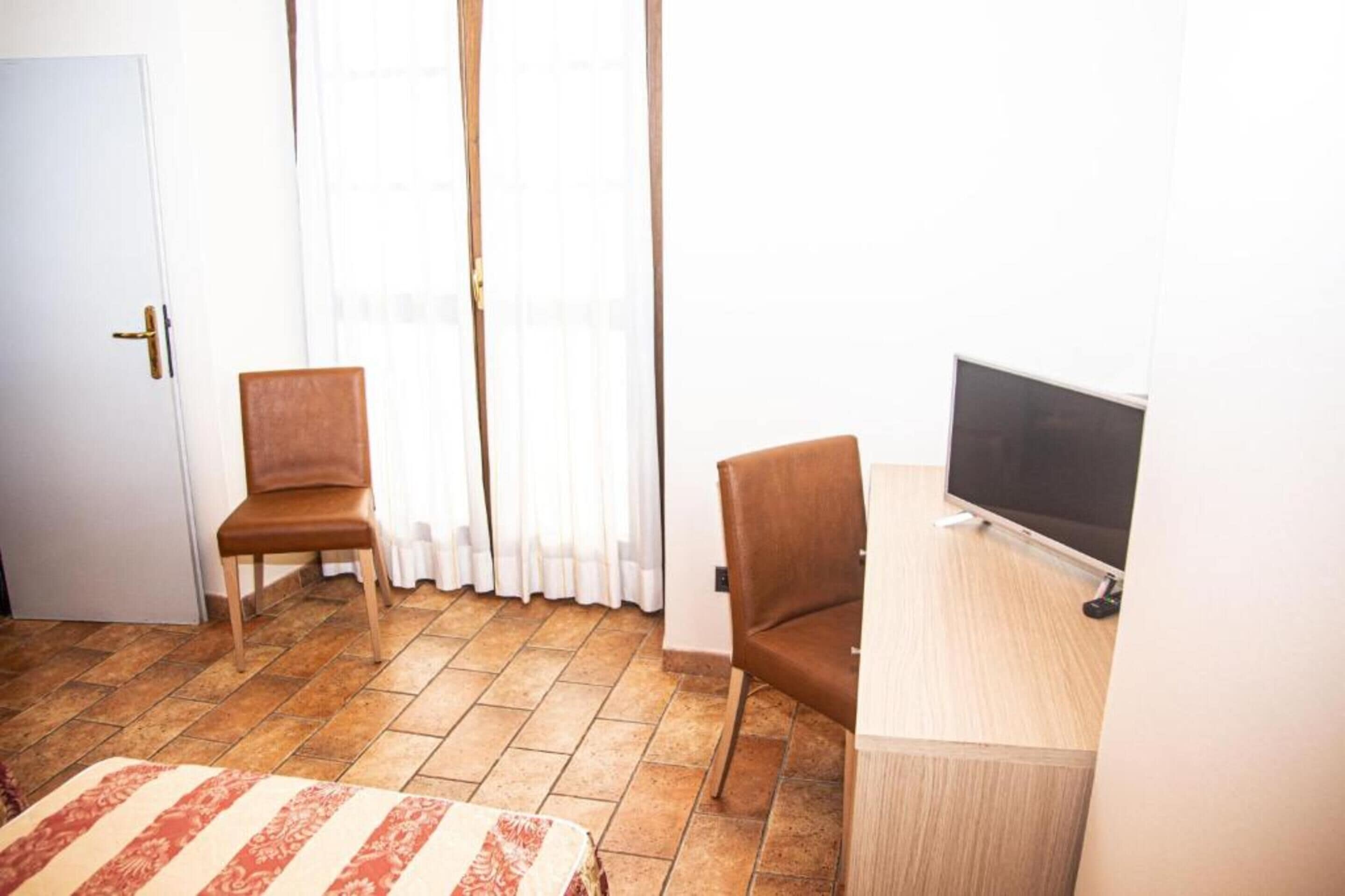 A room with a bed, two chairs, a desk with a TV, and a door.