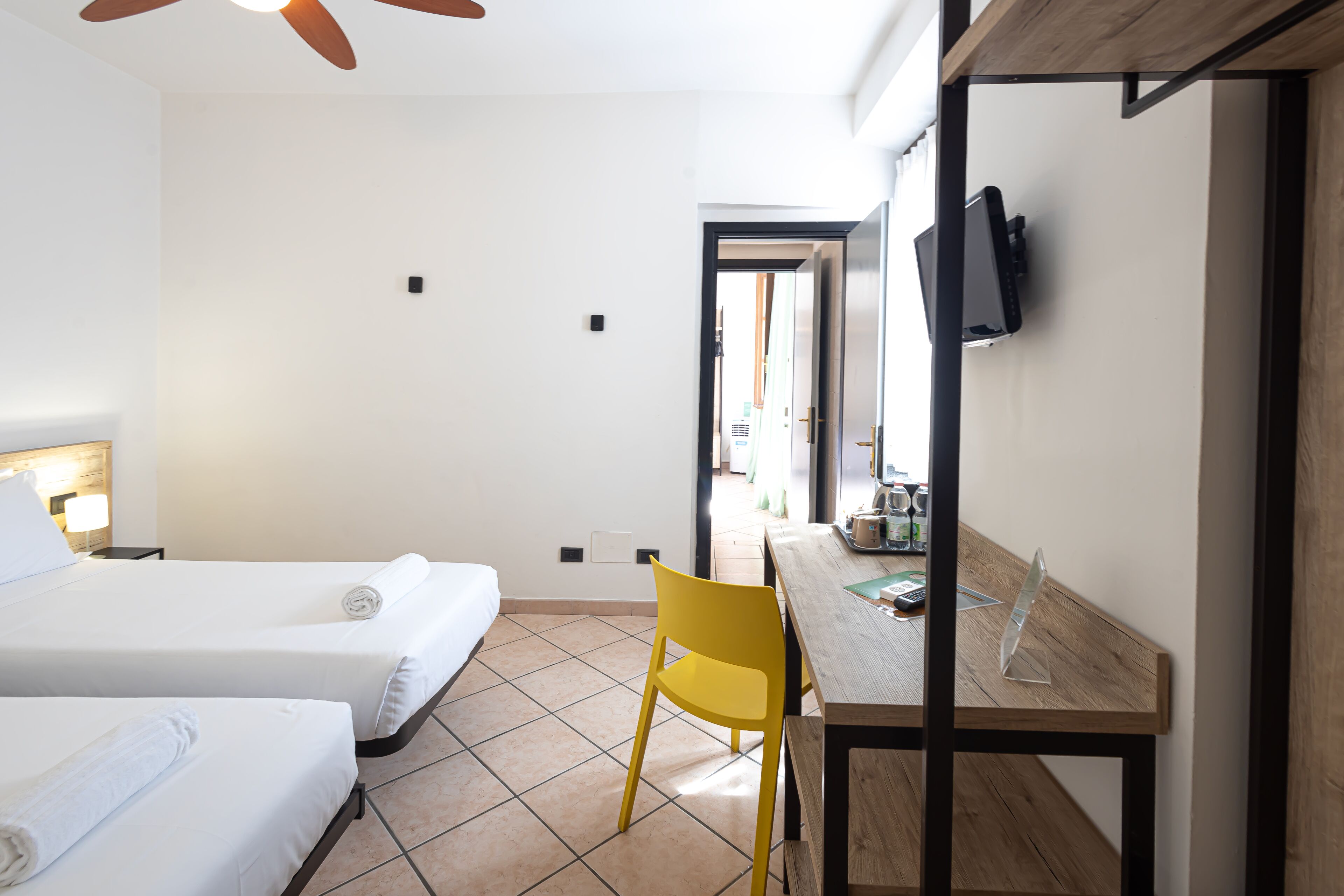 A hotel room with two beds, a wooden desk, a yellow chair, a TV mounted on the wall, and a visible door leading to another room.