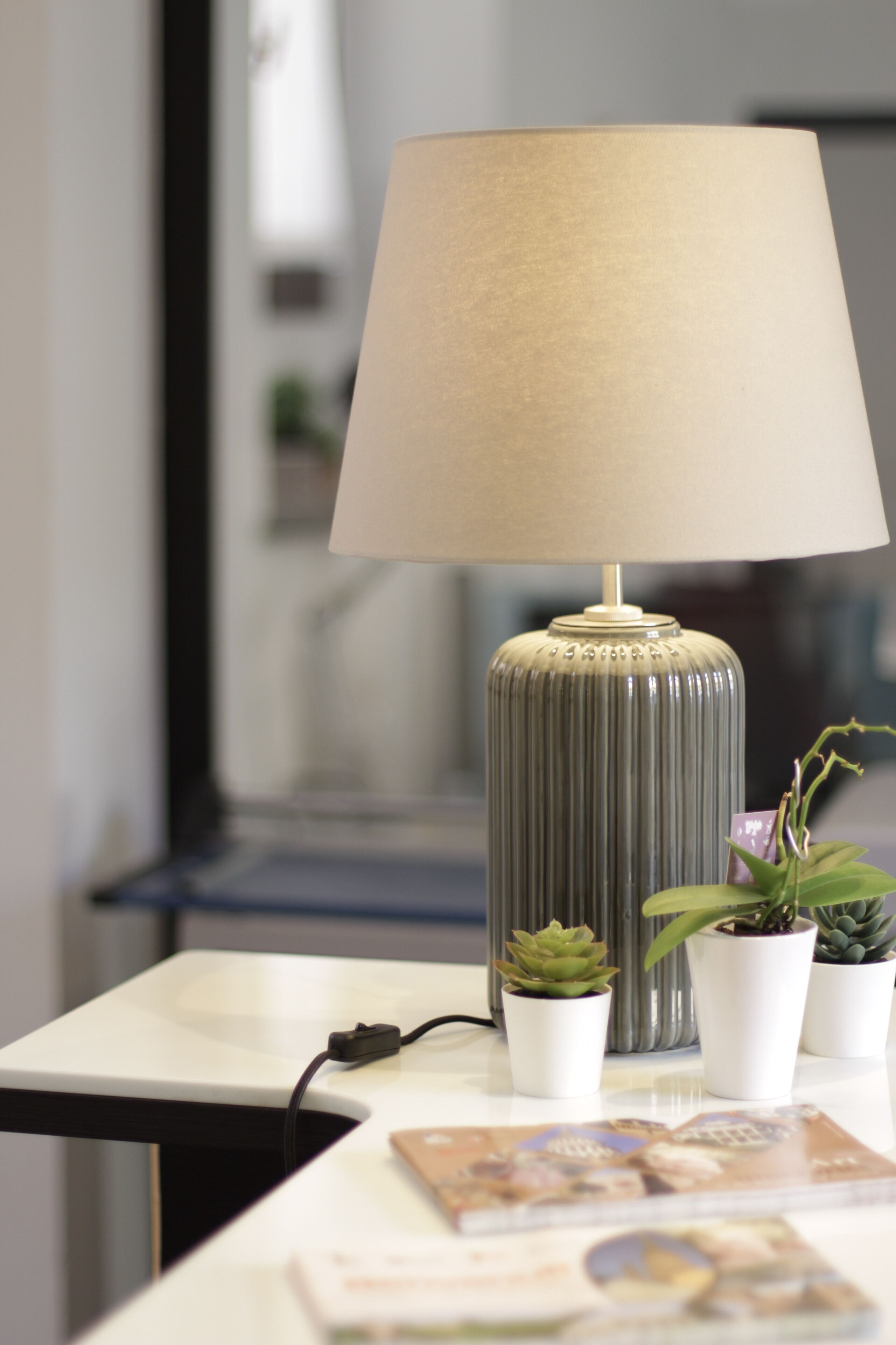 A lamp with a beige shade on a table, accompanied by three potted plants and a magazine.