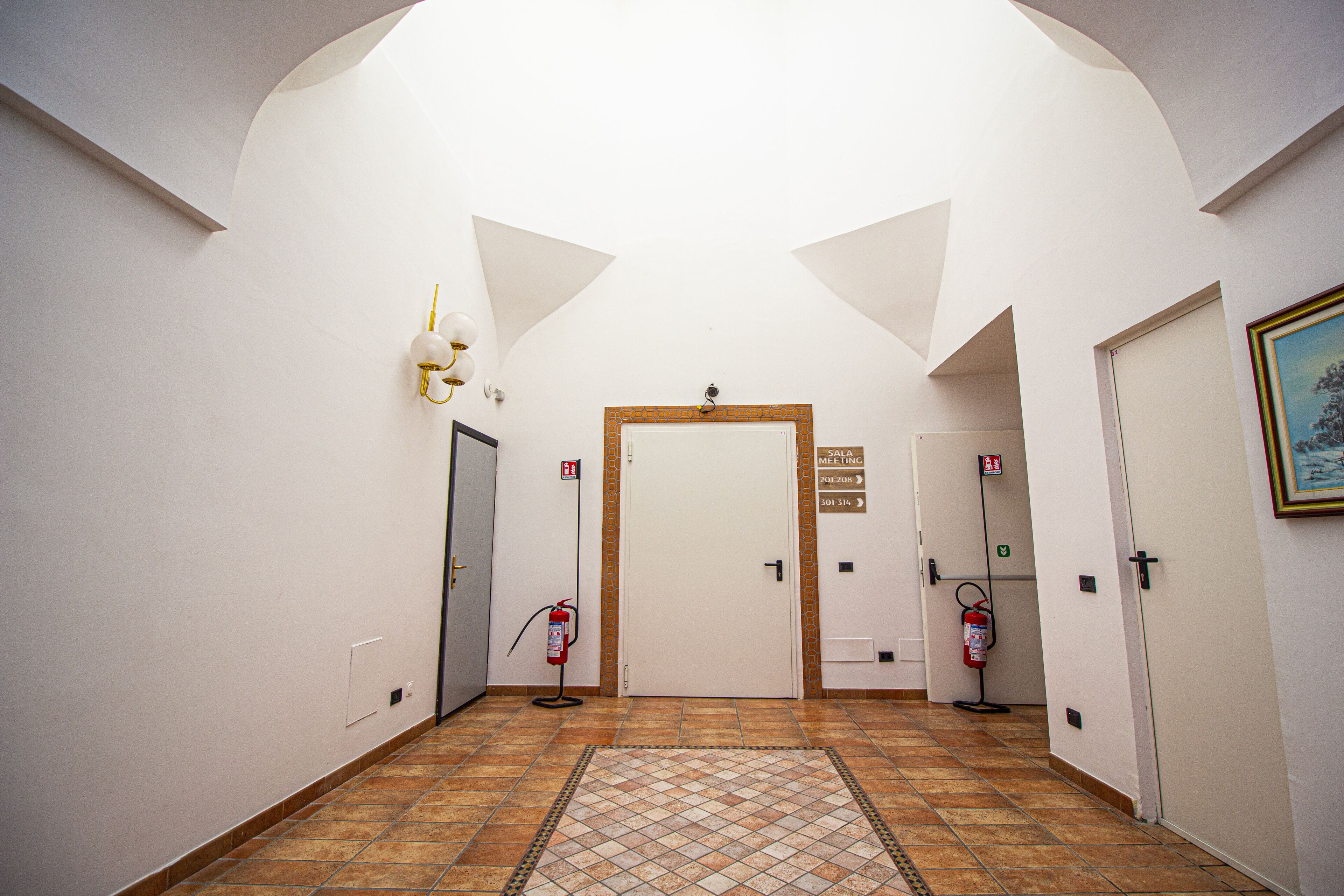 A hallway with a tiled floor, white walls, and a door at the end. There are two fire extinguishers mounted on the wall.