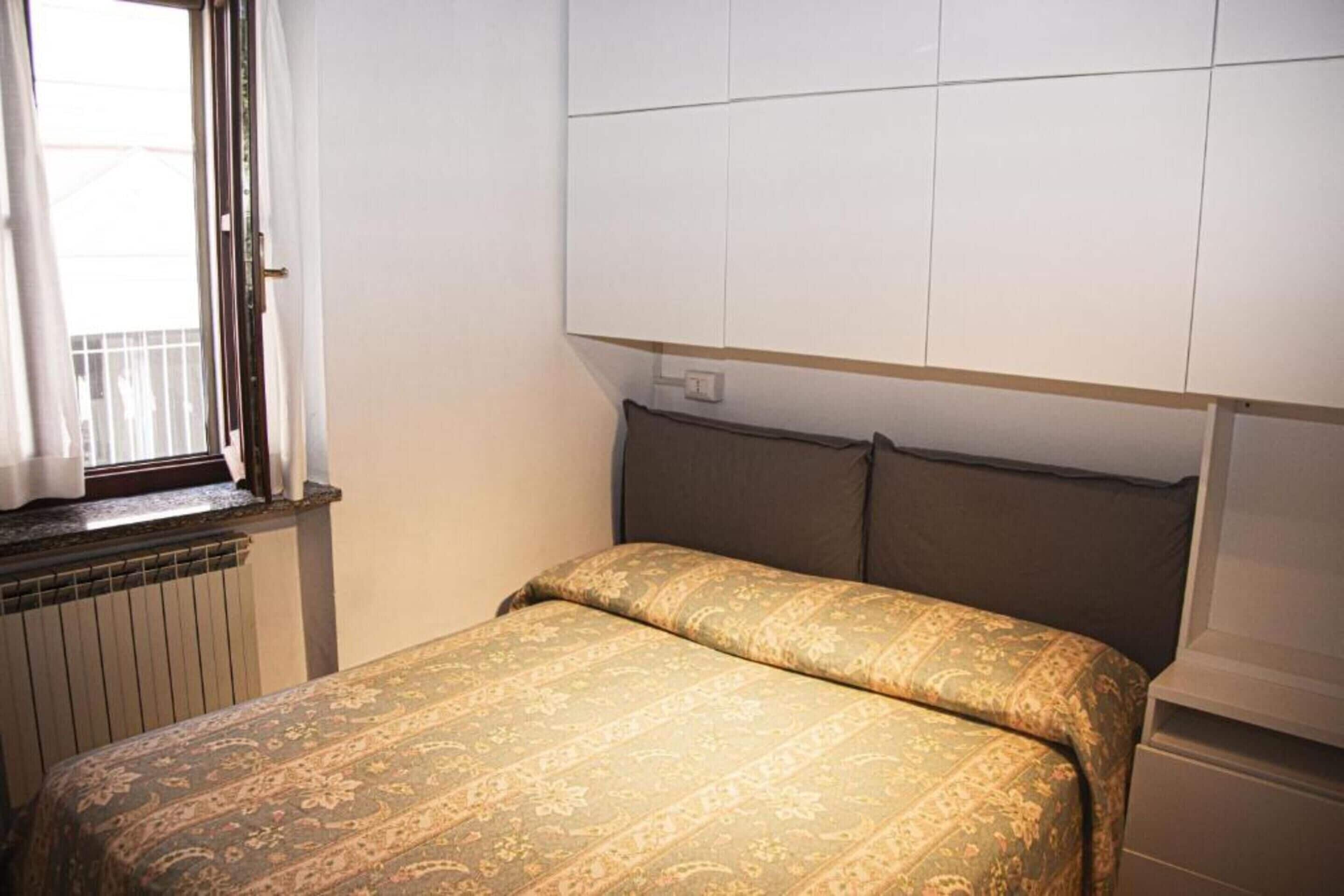 Un dormitorio con cama, ventana con cortinas, radiador y un mueble de pared.