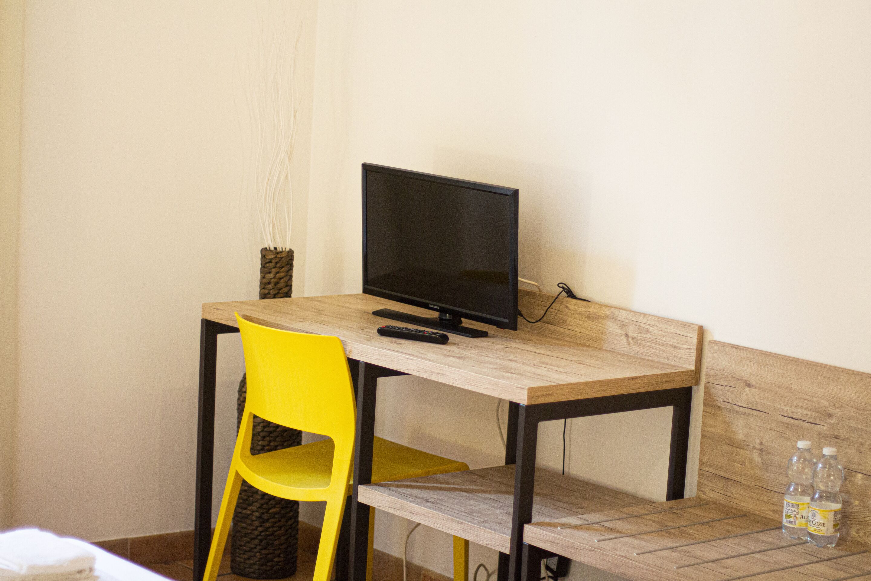 A small wooden desk with a black monitor, a yellow chair, and two bottles of water on the side.