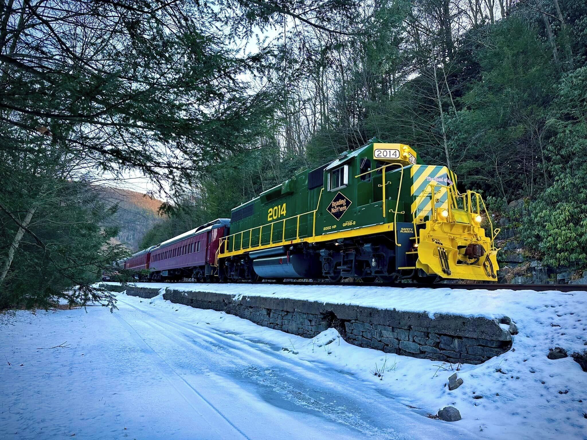 A green and yellow train with the number 2014 on the front, traveling through a snowy area with trees on either side.
