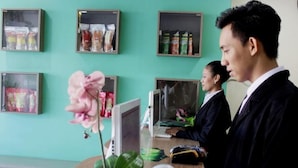 Un bureau avec un ordinateur, une plante en pot et un clavier. On y voit des étagères avec des bocaux et une fleur rose.