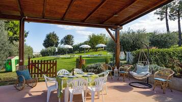 Eine Terrasse im Freien mit einer Holzpergola, weißen Kunststoffstühlen, einem für das Essen gedeckten Tisch und Blick auf eine Rasenfläche mit Sonnenschirmen und Stühlen im Hintergrund.