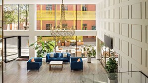 Een ruime lobby met een modern ontwerp, voorzien van een zithoek met banken en een salontafel, een trap en uitzicht op een geel gebouw met balkons.