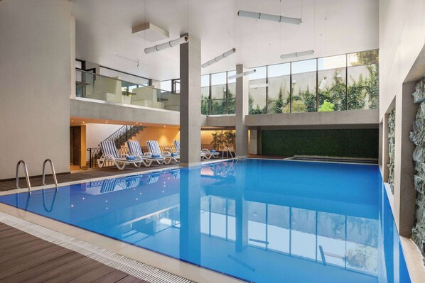 An indoor swimming pool with clear blue water, surrounded by lounge chairs and a staircase leading to an upper level. - Ramada by Wyndham Tbilisi Old City (Tbilisi)