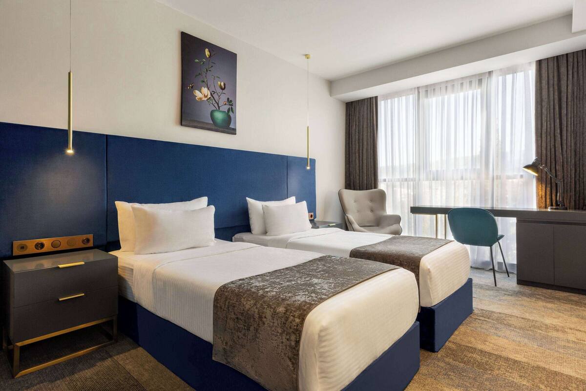 A hotel room with two beds, a desk, and a chair. There is a painting on the wall and a window with curtains.