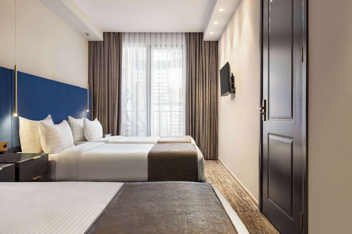 A hotel room with two beds, a large window with curtains, a flat-screen TV on the wall, and a dark door.