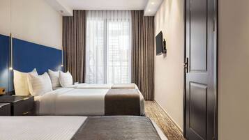 Una habitación de hotel con dos camas, un ventanal con cortinas, un televisor de pantalla plana en la pared y una puerta oscura.