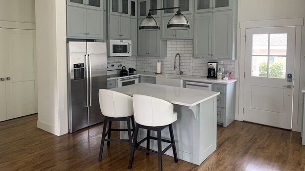 Une cuisine moderne avec des meubles gris, des appareils en acier inoxydable, un îlot central doté de deux tabourets de bar et un éclairage suspendu.