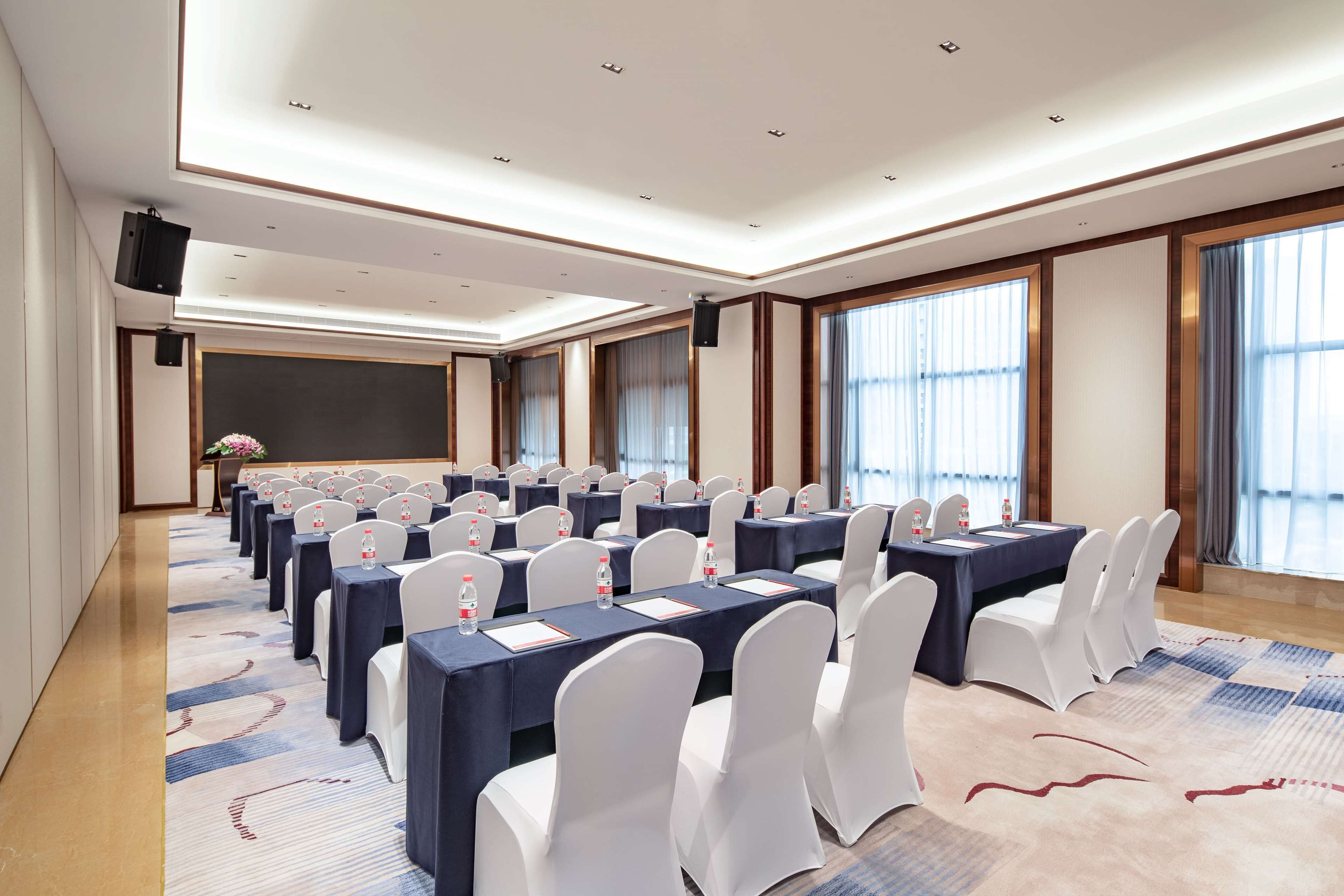 A conference room with rows of tables and chairs, large windows, and a carpeted floor.