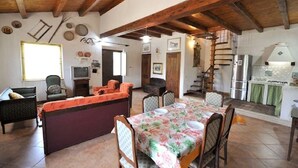 Un coin repas rustique avec une table en bois dressée pour quatre personnes, entourée de chaises. On aperçoit un canapé et un téléviseur à l’arrière-plan.