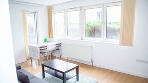 A bright living room with a sofa, a coffee table, and a dining area with a table and chairs. There are large windows with curtains and a radiator under the windows. - Large Shoreditch, Hoxton 3-bed Garden Flat (London)