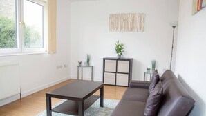 A living room with a dark brown sofa, a glass coffee table, a small side table with a plant, and a cabinet with a plant on top. - Large Shoreditch, Hoxton 3-bed Garden Flat (London)