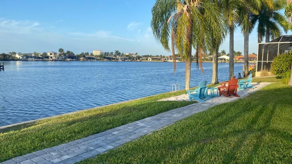 Una propiedad frente al mar con un paseo pavimentado, palmeras y mobiliario exterior junto al agua.