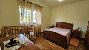A bedroom with a wooden bed, a wooden crib, a bedside table with a lamp, and a window with curtains.