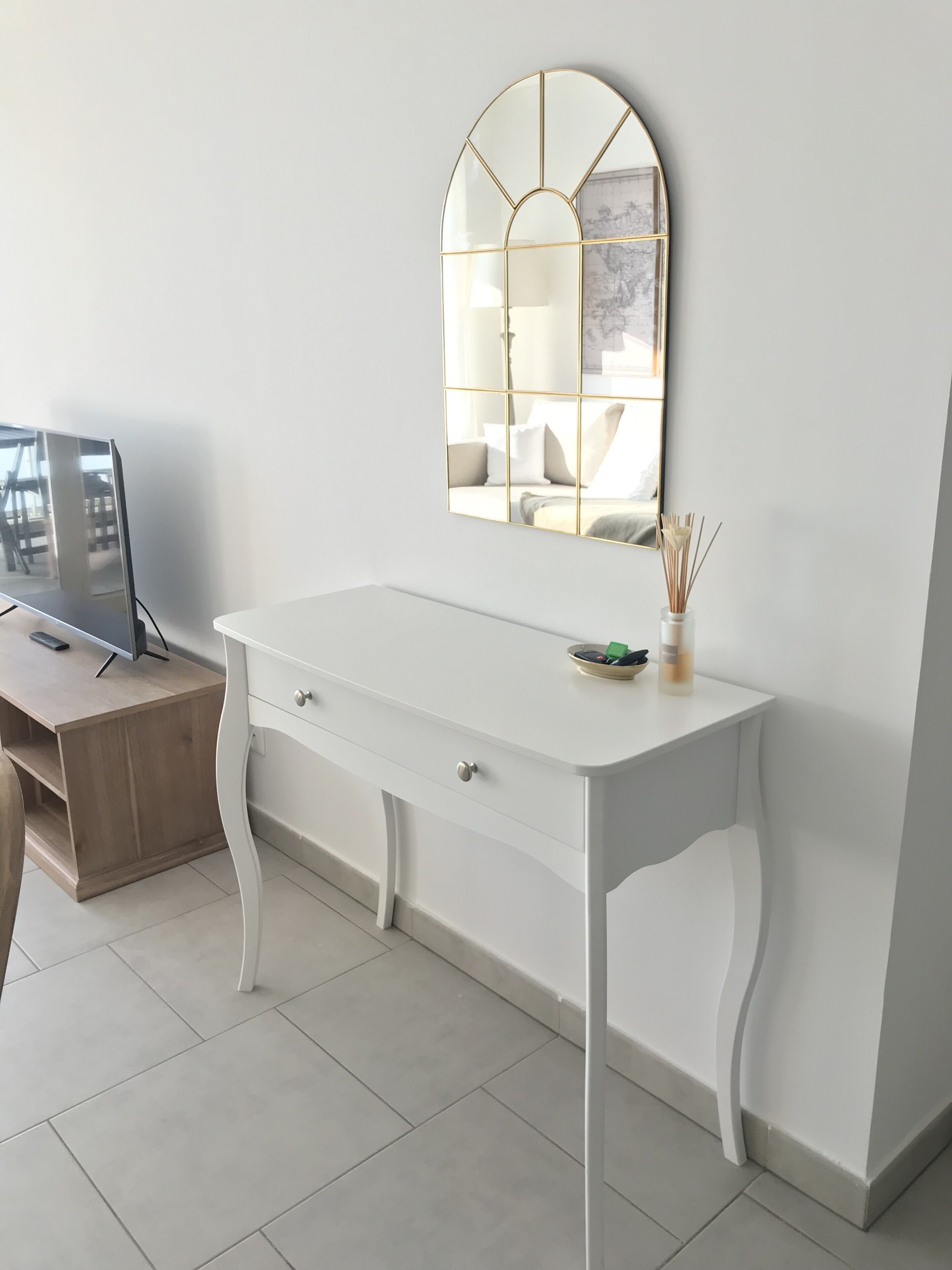 A white desk with drawers, a mirror with a gold frame, and a small table with a vase of reeds.