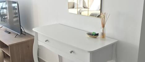 A white desk with drawers, a mirror with a gold frame, and a small table with a vase of reeds.