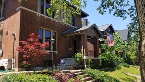 A two-story brick house with a front porch, a small garden with various plants, and an American flag.
