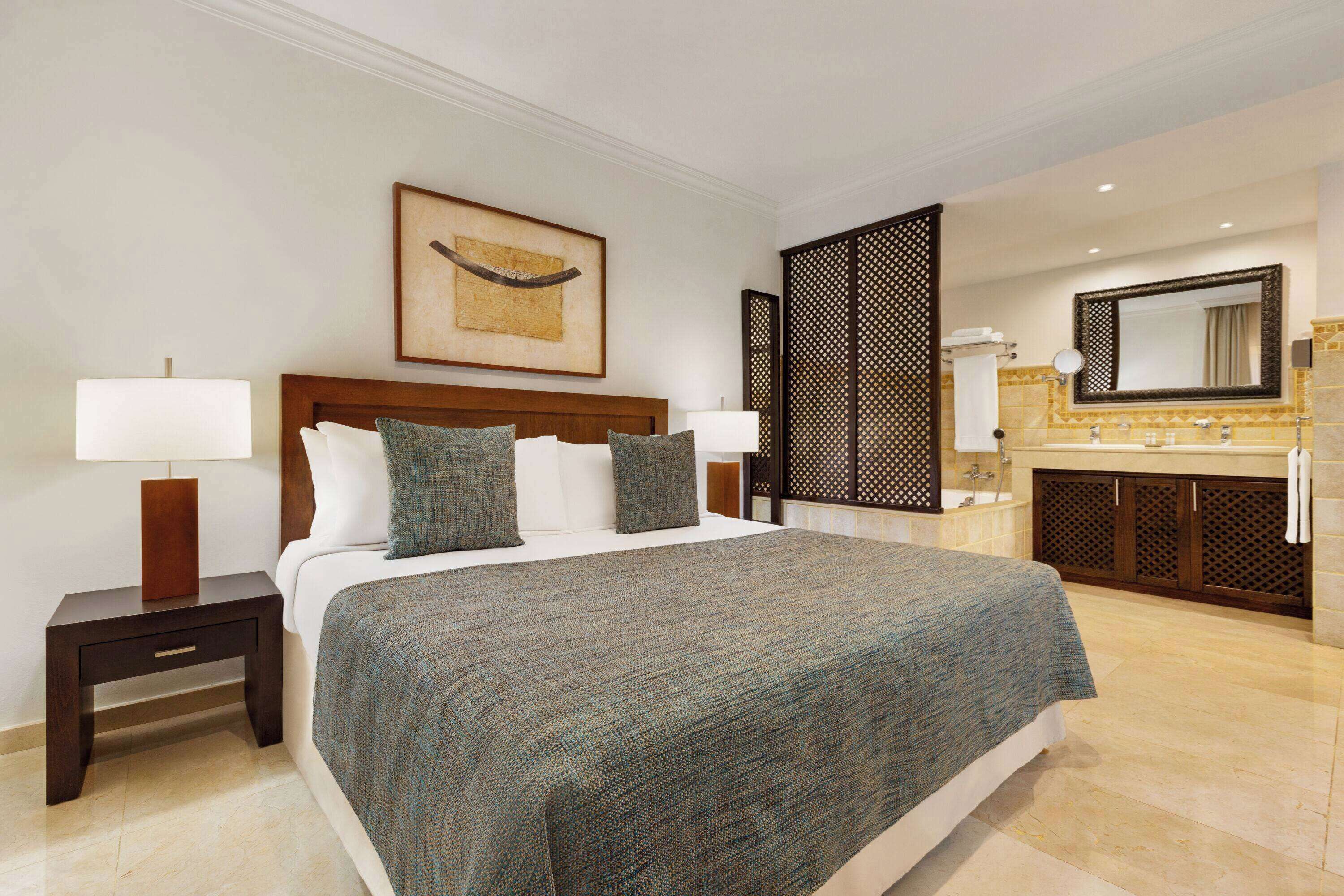 A hotel room with a large bed, a nightstand with a lamp, a bathroom with a sink and mirror, and a decorative wall piece.