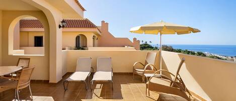 A terrace with a dining table and chairs, a yellow umbrella, and a view of the ocean.