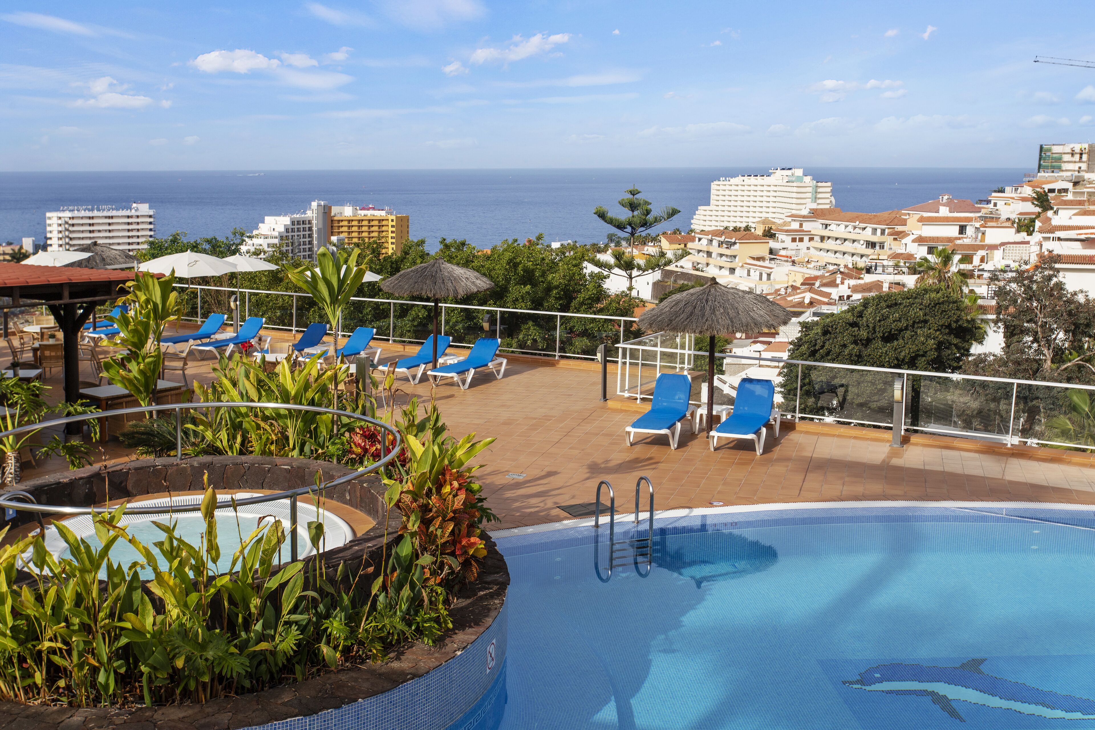 Un área de piscina en la azotea con sillones reclinables azules, un jacuzzi y vistas al océano y al paisaje urbano.
