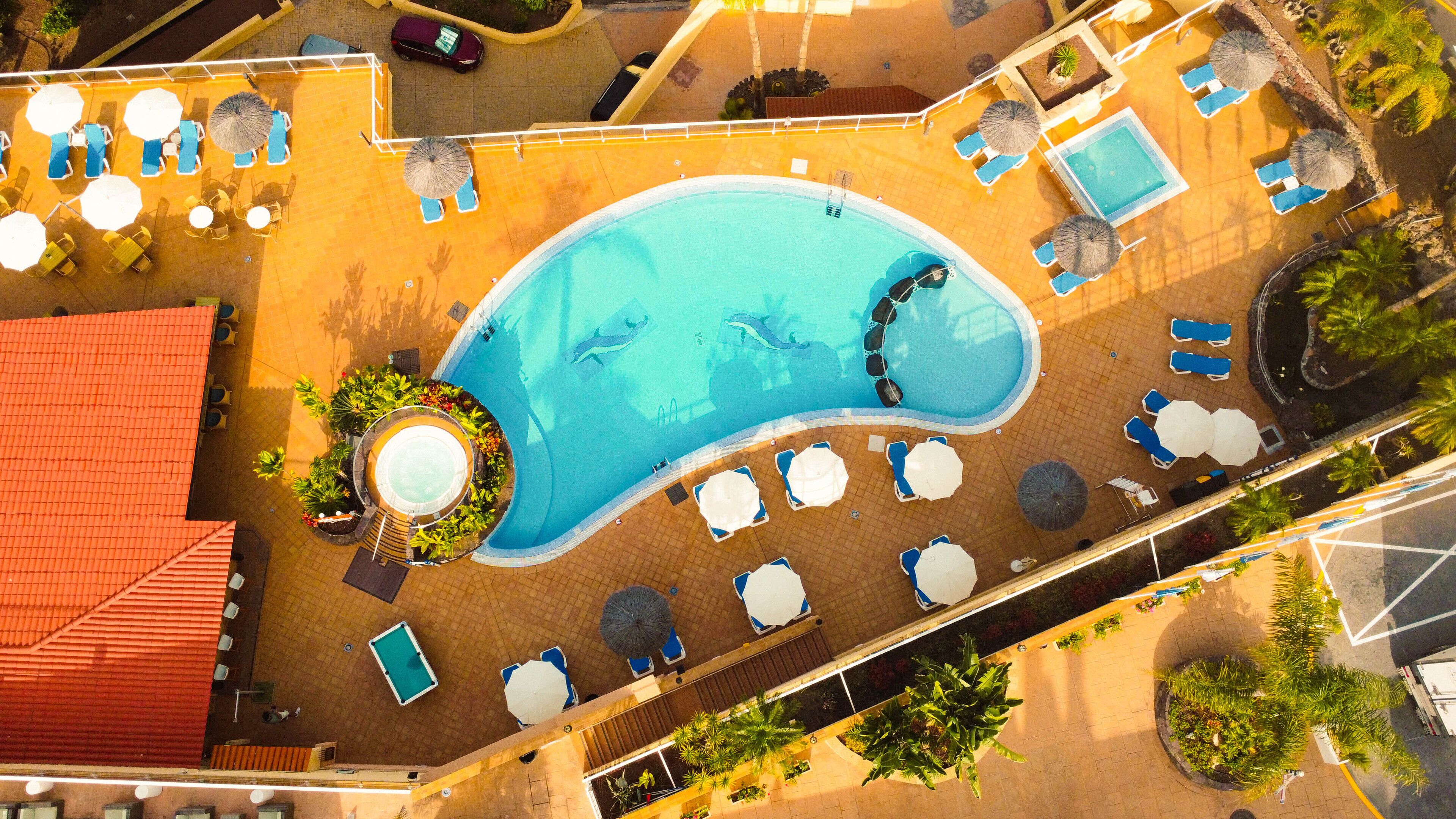 Vista aèria d'un hotel amb una gran piscina, diverses zones d'estar amb ombrel·les i un edifici amb teulada vermella.