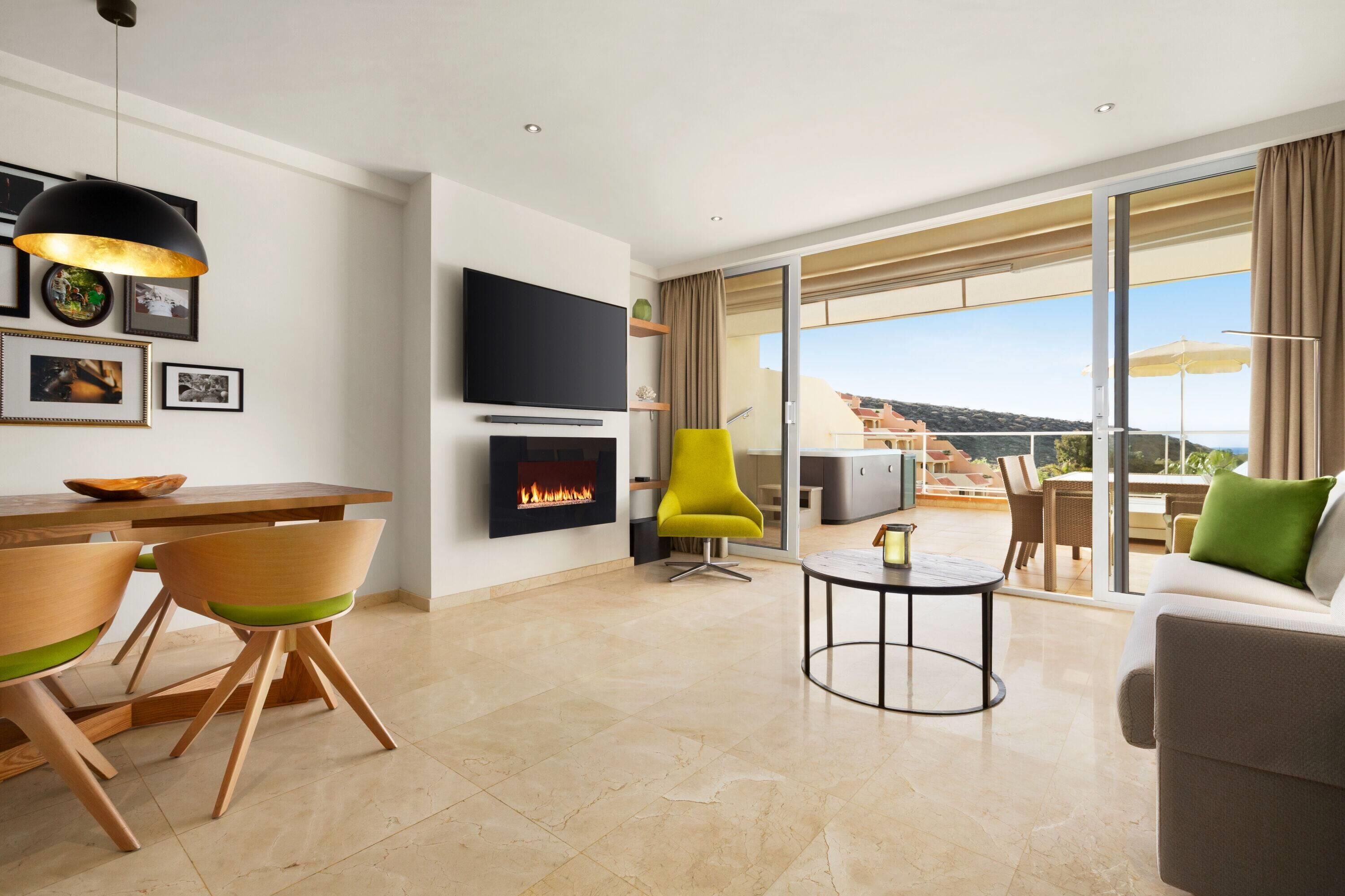 A modern living room with a fireplace, a flat-screen TV, a dining area with wooden chairs, and a balcony with a view.
