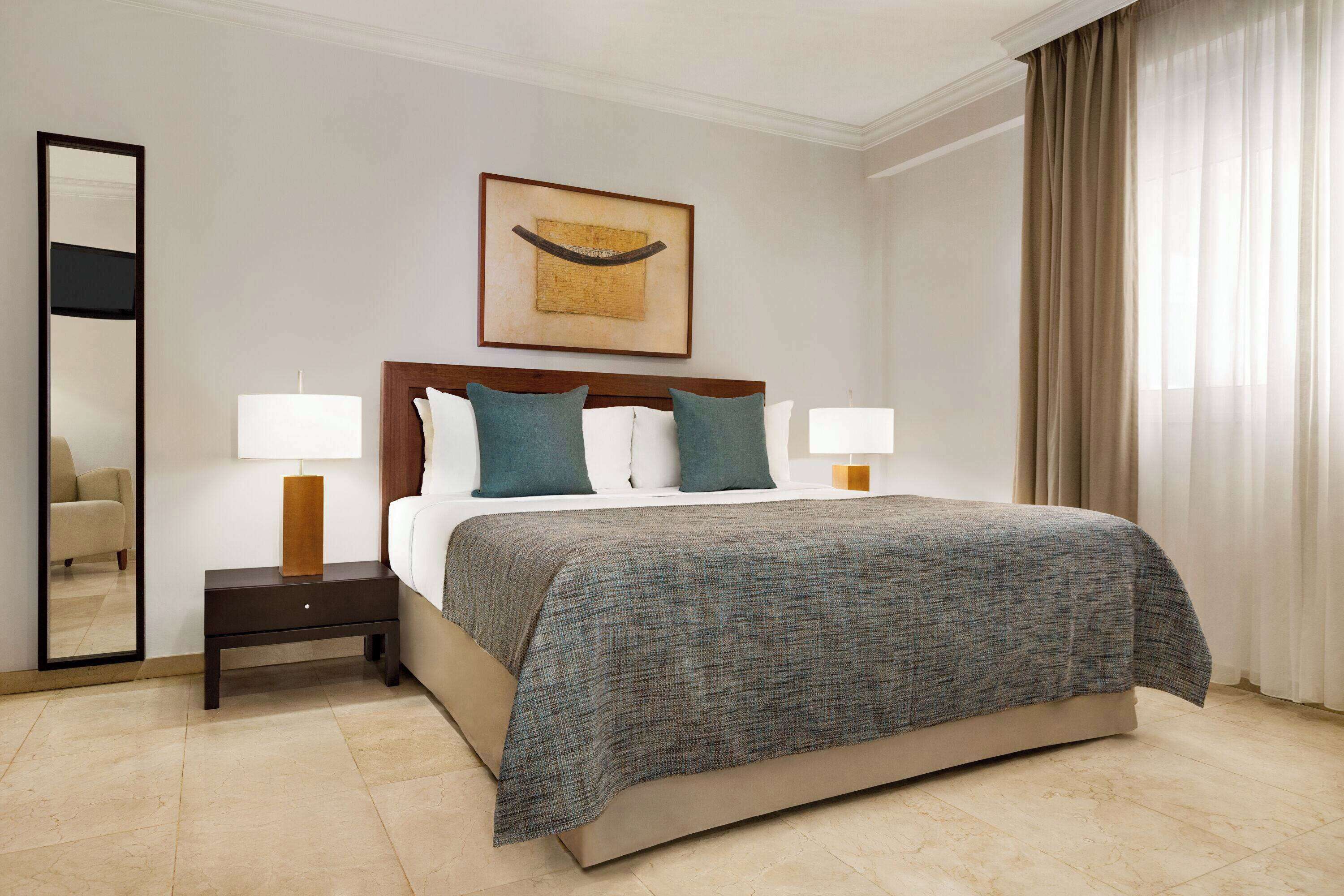 A hotel room with a large bed, two bedside lamps, a nightstand, a mirror, and a framed artwork on the wall.