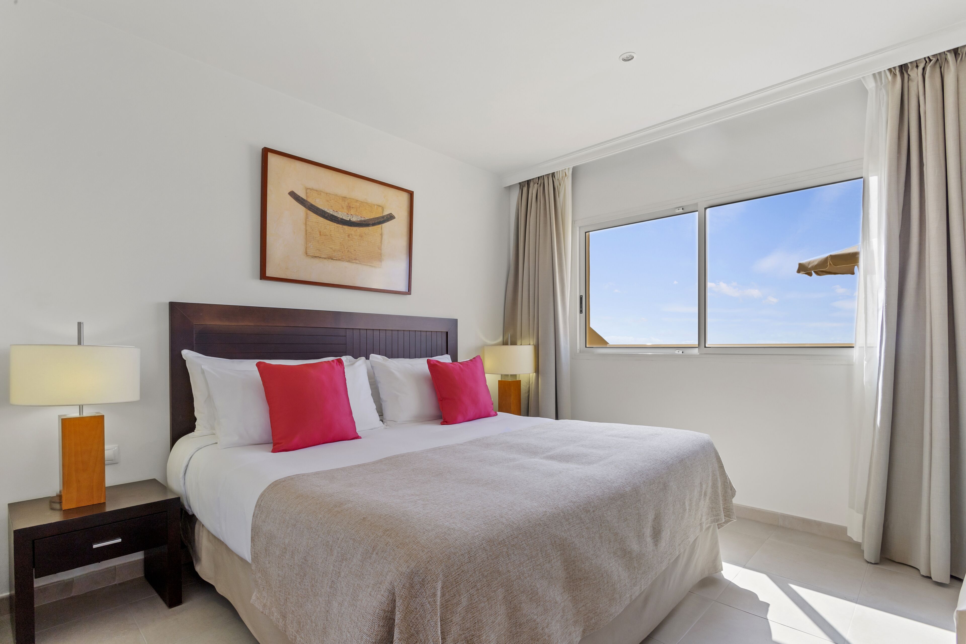 A hotel room with a large bed, two bedside tables with lamps, a framed artwork above the bed, and a window with curtains.