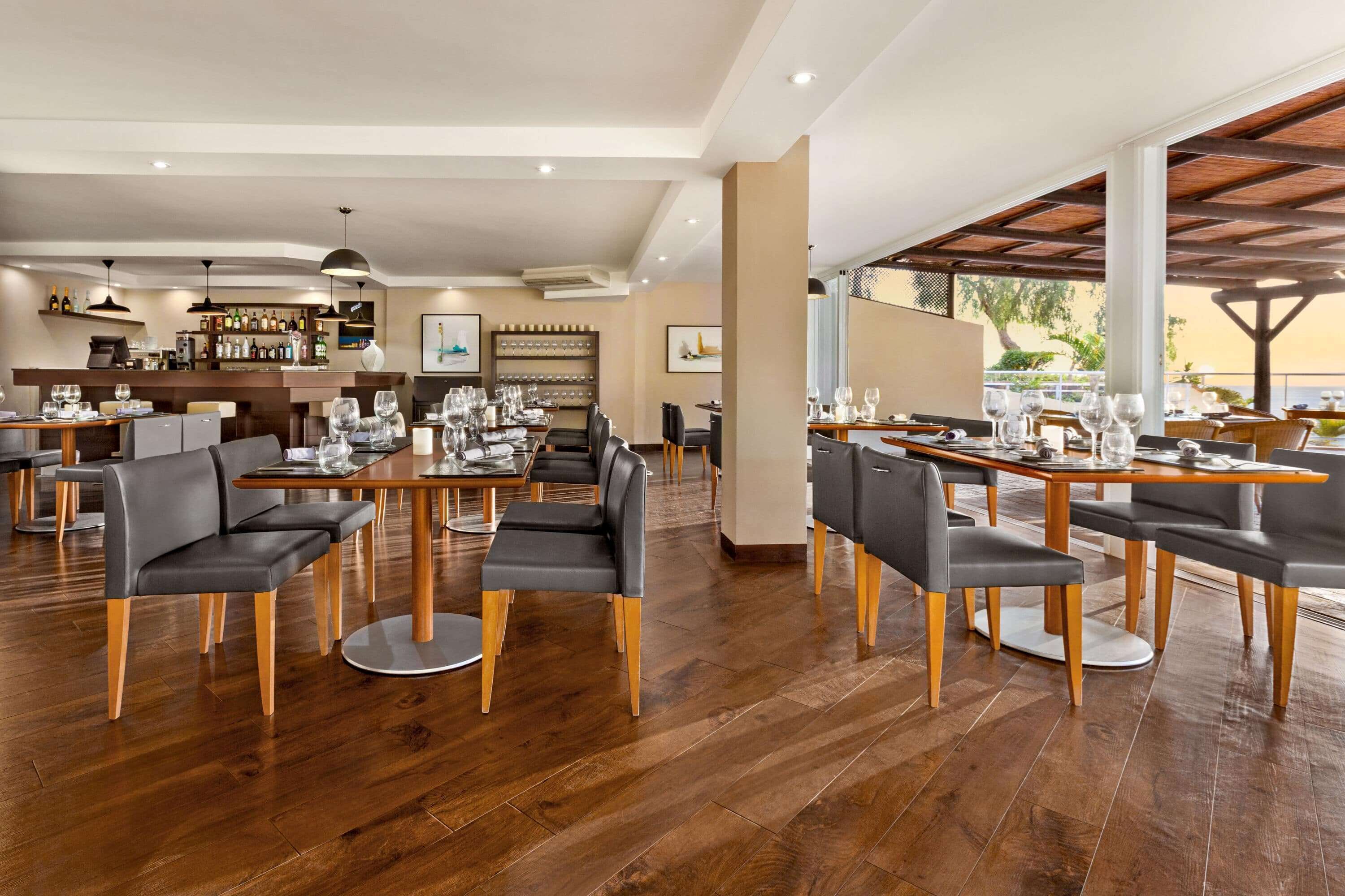 A modern restaurant with wooden flooring, black upholstered chairs, and a bar area with wine bottles.