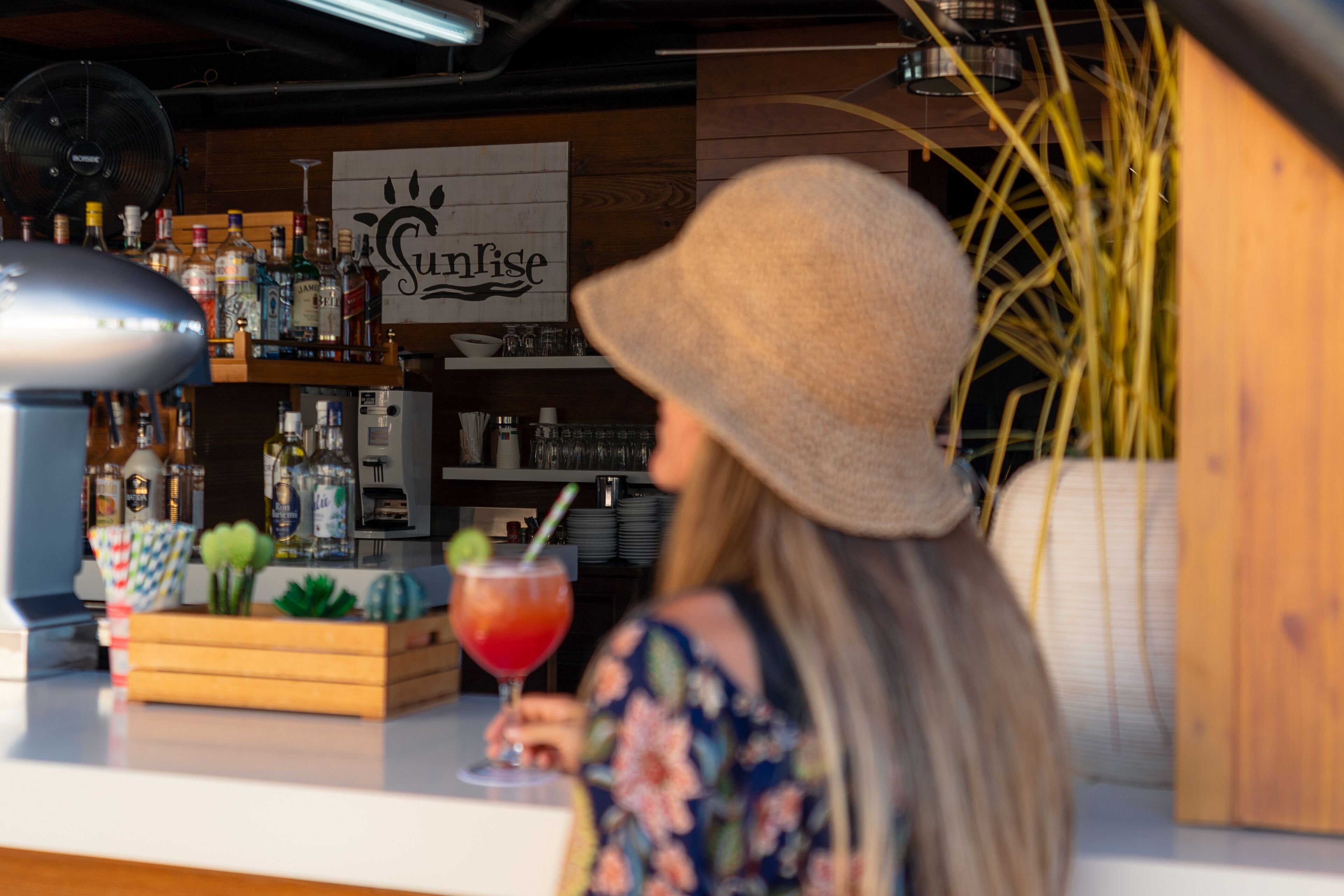 Une femme portant un chapeau est assise au bar. On peut voir une enseigne avec le mot « Sunrise » et diverses bouteilles et verres disposés sur des étagères.