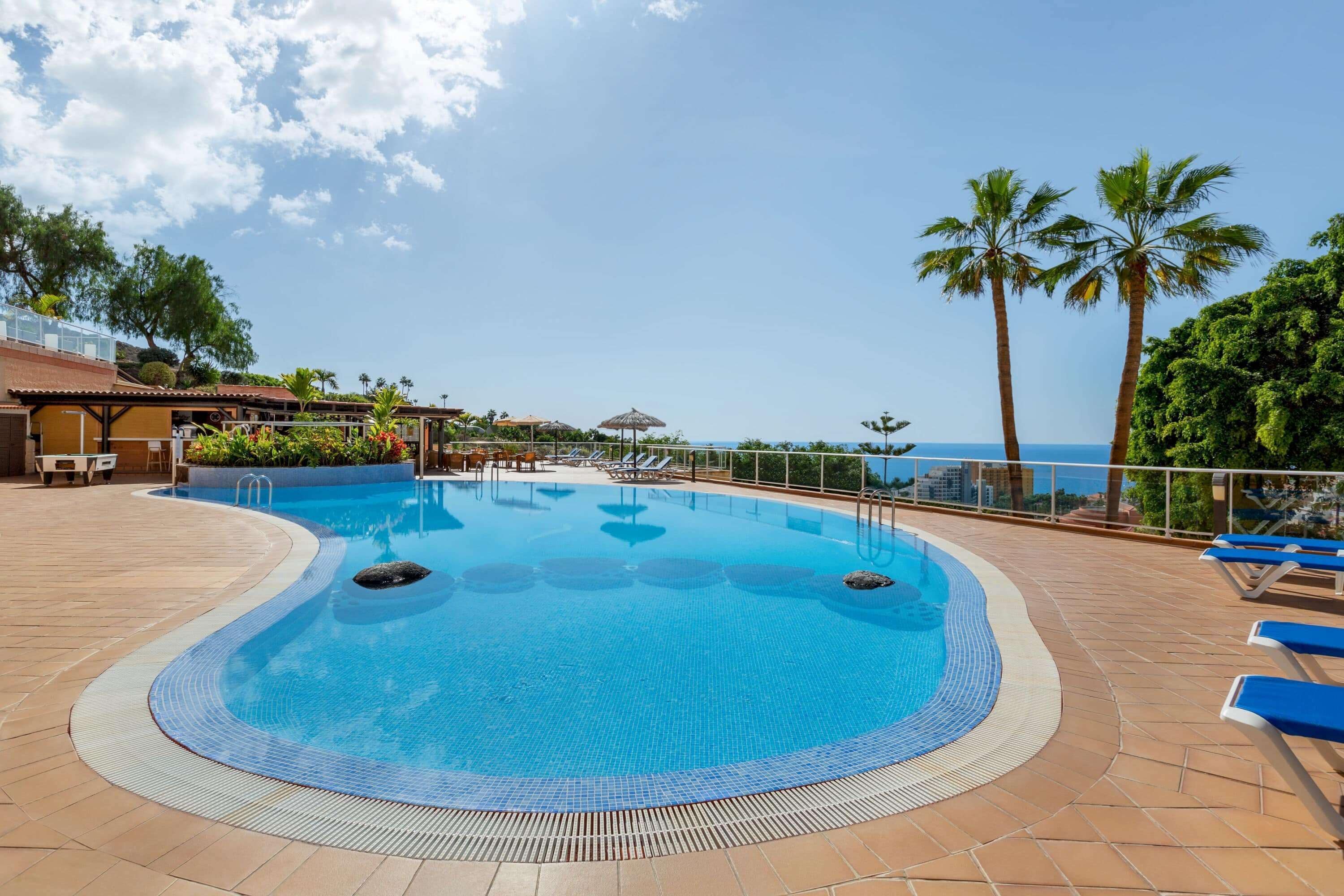 Ein Pool mit geschwungener Kante, umgeben von Liegestühlen und Palmen, mit Blick auf das Meer im Hintergrund.