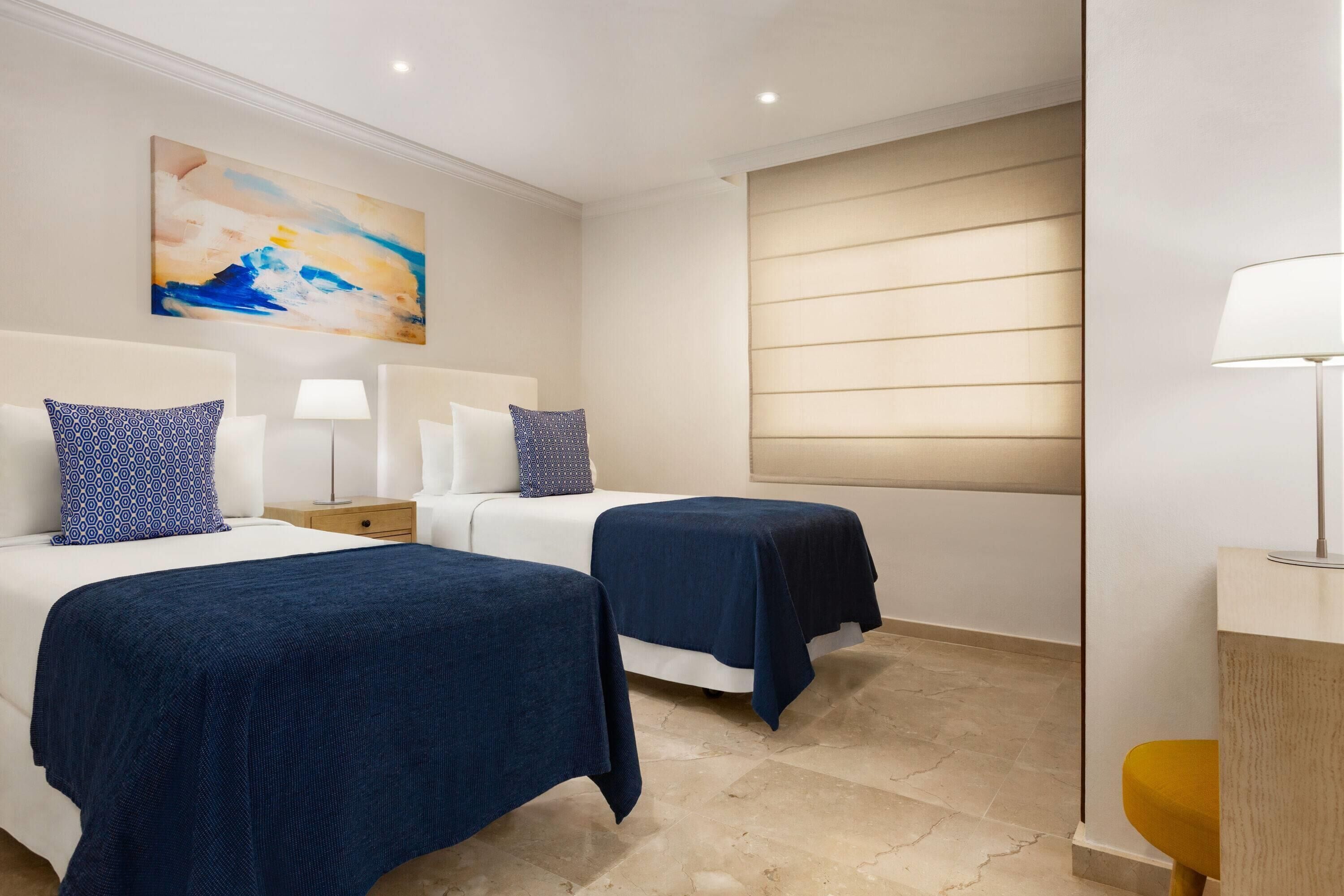 Hotelkamer met twee bedden, een nachtkastje, een lamp, een schilderij aan de muur en een raam met jaloezieën.