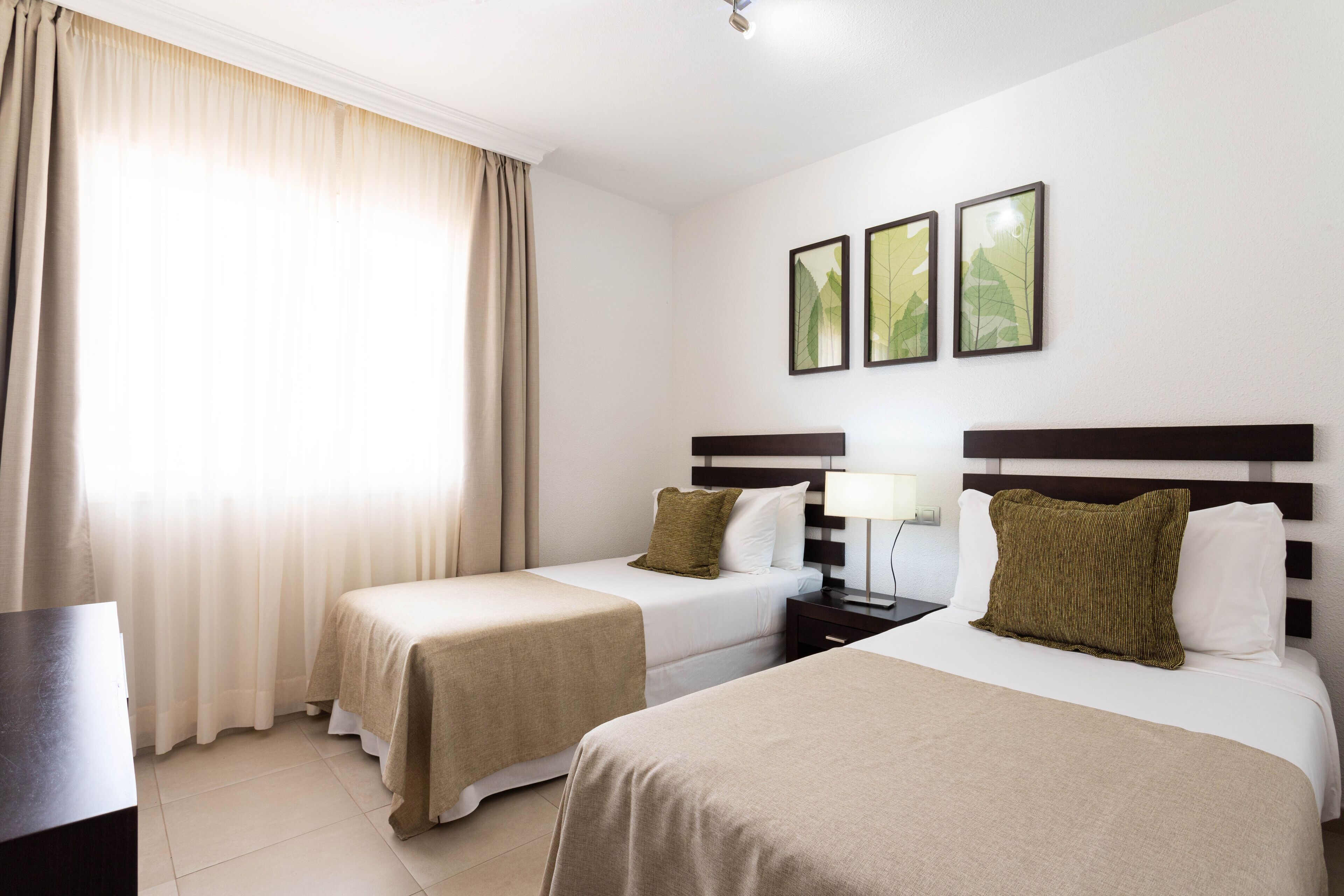 A hotel room with two beds, a nightstand, a lamp, and a window with curtains.