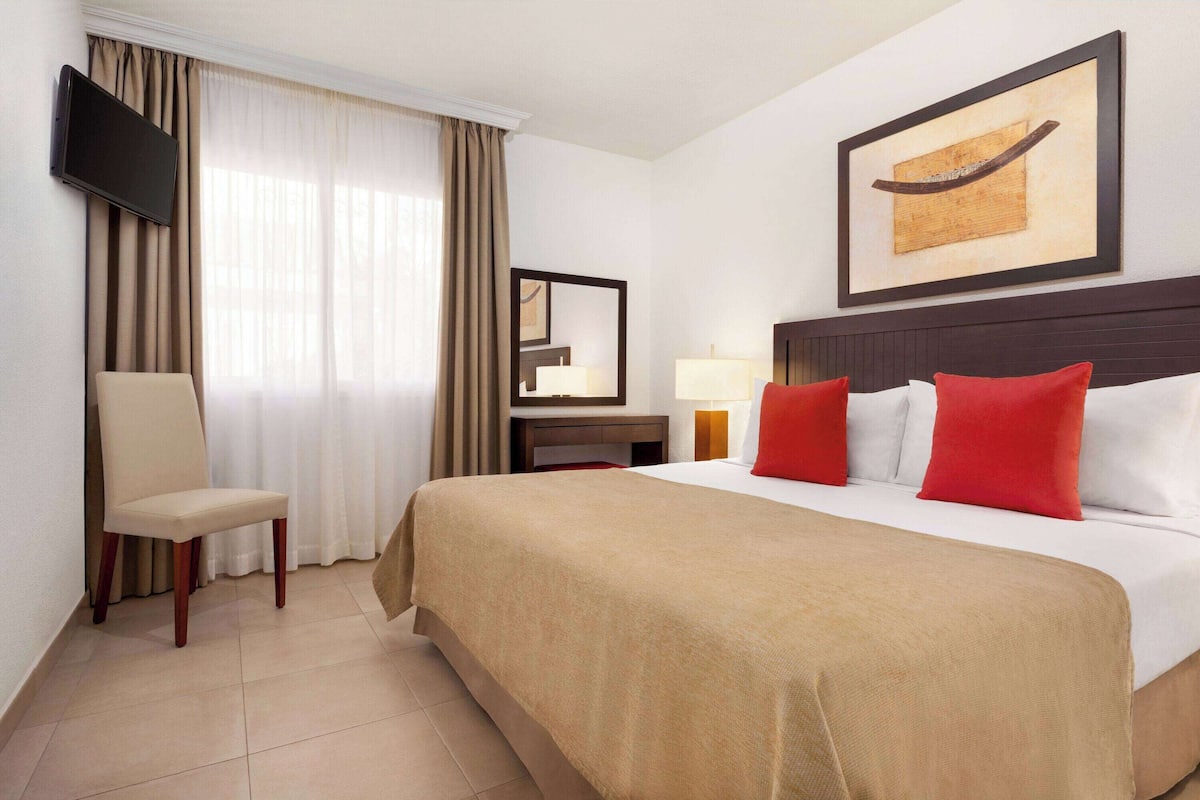 A hotel room with a bed, a chair, a desk with a lamp, a television, and a painting on the wall.