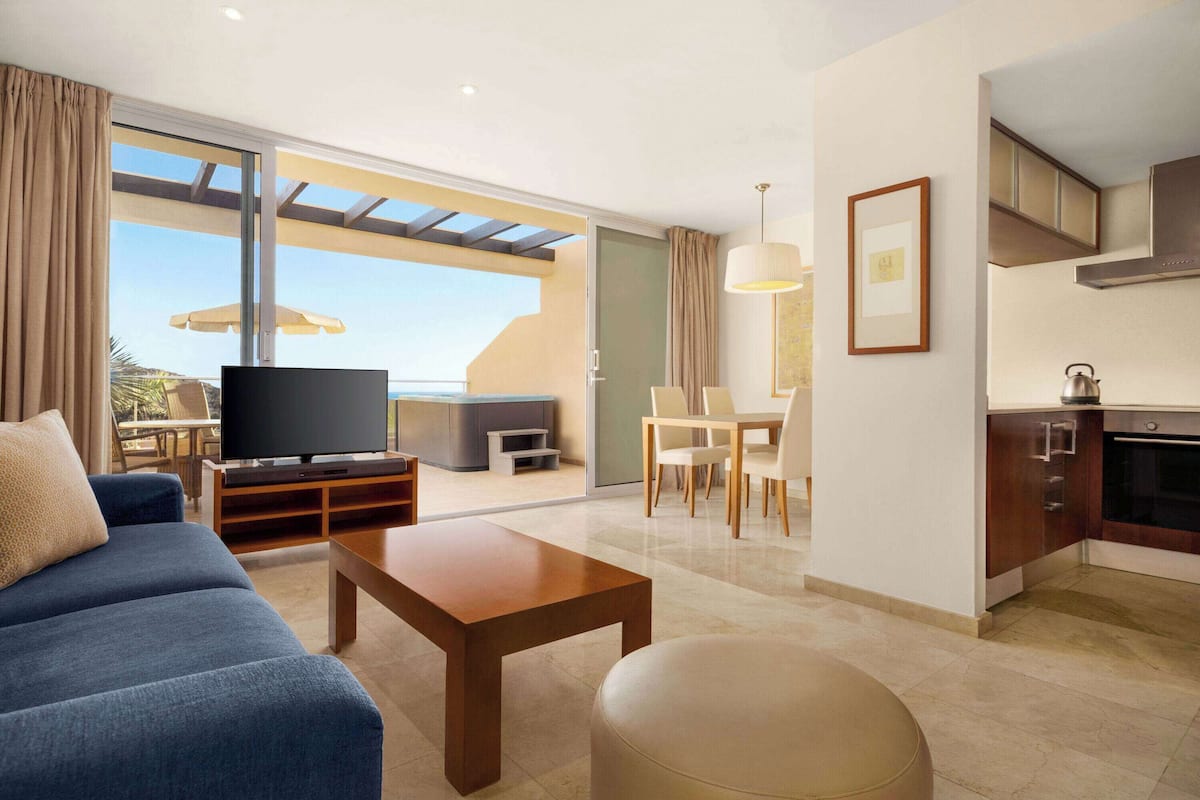 A modern living room with a sofa, coffee table, and TV, a dining area with chairs, and a kitchen with cabinets and a stove.