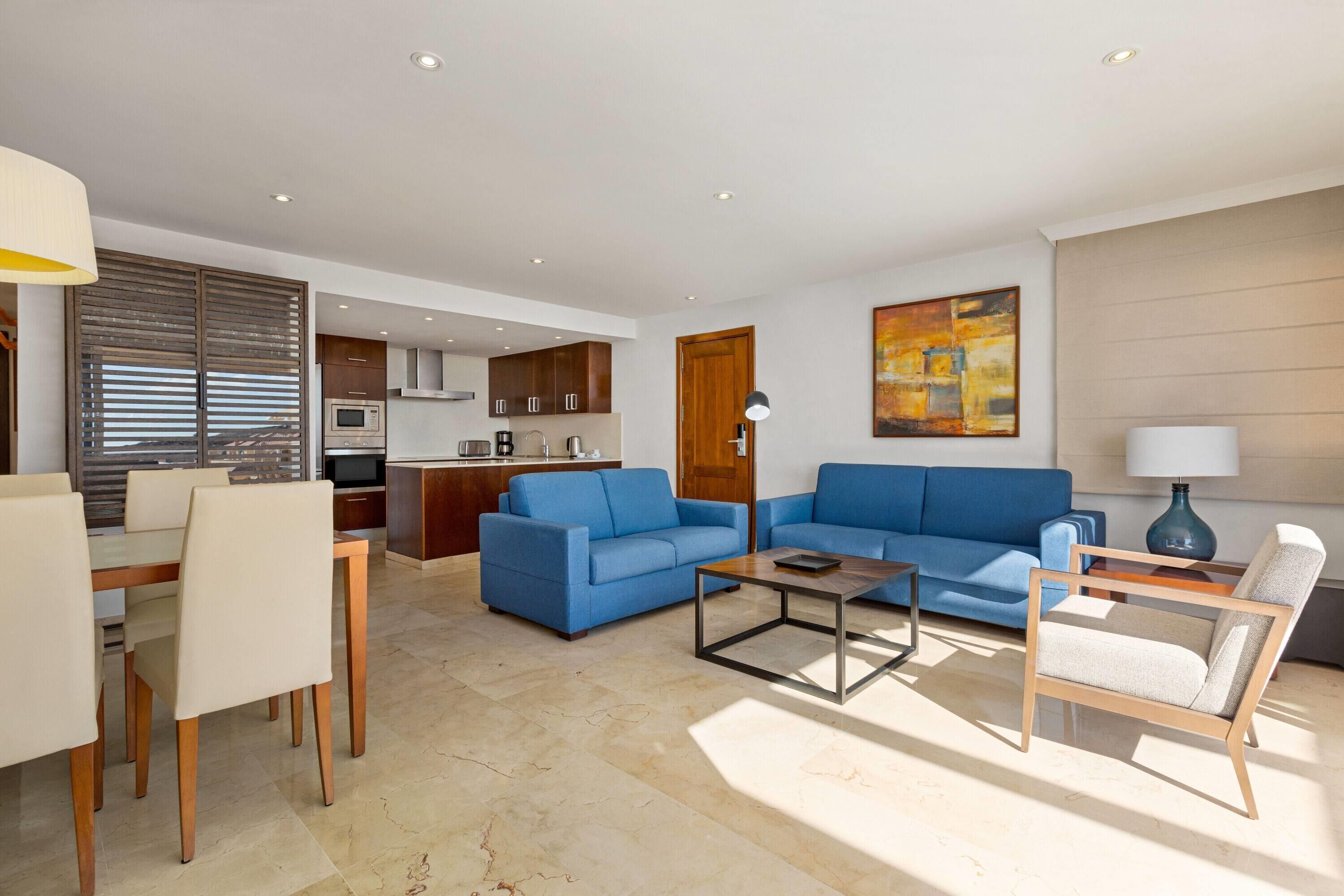 A modern living room with a blue sofa, a wooden coffee table, and a dining area with a table and chairs.